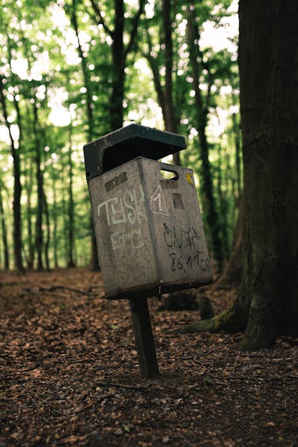 A weathered grey metal rubbish deposit box with a black open lid is mounted on a slightly tilted metal pole, situated on a woodland forest floor covered in brown, textured soil and dry leaves. The box exhibits graffiti tags and scribbles in black and white, indicating it has been vandalized. Surrounding the unit are tall tree trunks with dark, rough bark, and a canopy of green foliage allowing soft natural light to filter through, creating a dappled effect on the ground. The background features densely packed trees with blurred leaf patterns, contributing to the tranquil woods atmosphere. The scene suggests an outdoor setting where waste management or disposal is managed independently, possibly supporting local or private waste removal services. Waste Clearance Barnet might address the need for on-site clearance or private rubbish collection in such natural environments, aligning with alternative waste handling practices outside conventional urban waste systems. The image emphasizes the importance of maintaining clear and appropriate waste disposal points in public or semi-natural spaces to reduce illegal dumping or littering.