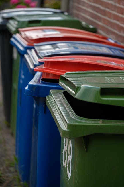 A row of six wheelie bins lined up along a pavement next to a brick wall, with their lids closed. The bins are arranged in a sequence of green, blue, red, blue, red, and green, from front to back. The green bin in the foreground has a partially visible recycling symbol painted in white on its side, and the lids are made of ribbed plastic with a smooth finish. The blue and red bins behind are similarly constructed, with the red lids appearing slightly curved and reflective under natural light. The background shows a residential setting with a motor vehicle partially visible, parked behind the bins, and the area appears clean and orderly. This scene is representative of private waste disposal arrangements that may be coordinated through local rubbish removal services, such as Waste Clearance Barnet, ensuring proper waste segregation and collection outside residential properties.