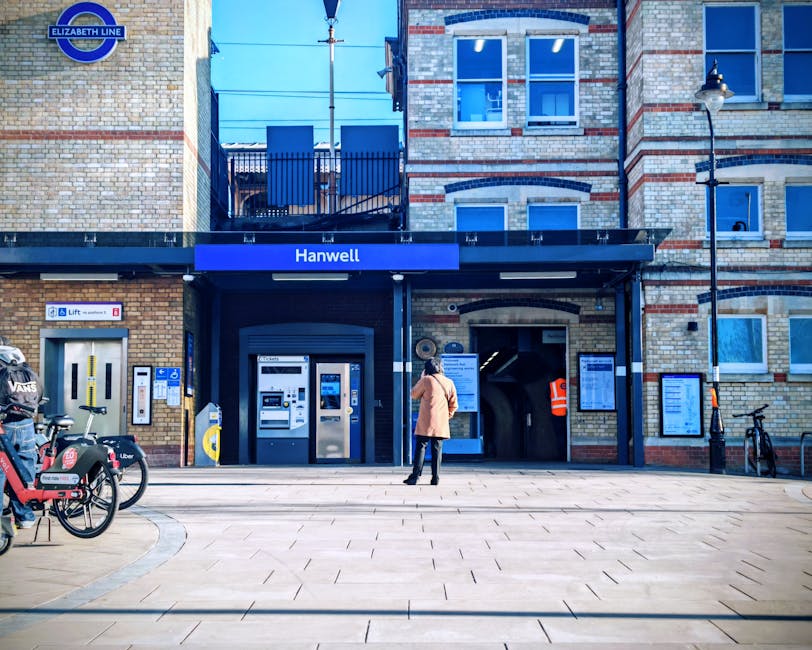 A woman in a beige coat and black trousers stands on a paved area in front of the entrance to Hanwell station, part of the Elizabeth Line. The station's facade features a brick exterior with a black canopy displaying a blue sign with white text that reads 'Hanwell.' To the left of the entrance, there are two white ticket machines and an automated payment kiosk. A yellow and grey card reader is mounted nearby, and a small sign indicates lift access for disabled passengers. On the left side of the image, a row of bicycles is parked on the pavement, including one with a child seat and another with a prominent red logo. To the right of the station entrance, there is a narrow, unlit underground access door, with informational signage mounted on the brick wall above it. A streetlamp with a classic design is attached to the right side of the building, extending over the sidewalk. The overall scene is lit naturally with clear weather, and the environment suggests a busy urban setting where independent, private waste disposal may occur alongside public transport services, fitting within a typical rubbish removal context.