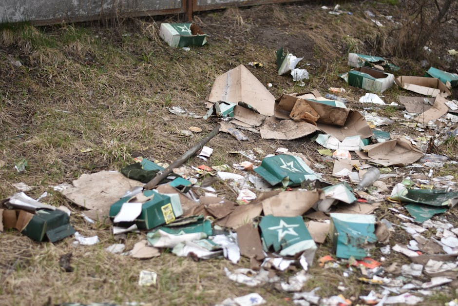 An outdoor scene showing a patch of ground near a grassy embankment, scattered with discarded cardboard boxes, many of which are torn and crumpled, with some displaying a green and white star logo. The boxes vary in size, some flattened while others remain partially intact, alongside loose paper and packaging debris. A metal pole, possibly part of a fence or equipment, lies diagonally across the space, partially obscured by the rubbish. The background includes a weathered wooden fence and sparse vegetation, indicating an unmanaged area. The lighting is natural, suggesting daytime with overcast weather, and the litter appears to have been randomly discarded, characteristic of unauthorised dumping. This visual context reflects the need for proper rubbish clearance, such as those provided by waste management services specializing in private waste removal or on-site clearance solutions, like those offered by Waste Clearance Barnet.