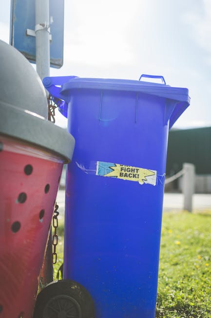 A large, upright blue recycling bin with a partially open lid is positioned on a grassy area outdoors, with a small yellow and blue label reading 'FIGHT BACK!' taped across its front. To the left of the bin, part of a pink rubbish or recycling container with a grey lid and black dotted pattern is visible, with a short chain attached to the side. In the background, there is a metal pole, a blue sign or board, and a road with guardrails, suggesting a roadside or driveway setting. The scene is lit by natural daylight, with some clouds visible in the sky, indicating an outdoor environment suitable for private waste collection or rubbish removal services conducted by Waste Clearance Barnet, focusing on efficient and safe rubbish handling in the local area.