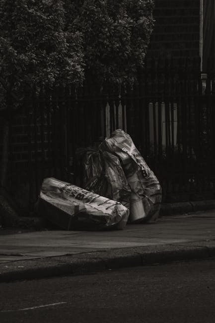 Three large black rubbish bags made of plastic, tightly tied at the top, are placed on a paved sidewalk near the edge of a road. The bags appear to contain miscellaneous waste materials and are irregularly shaped, with some leaning or slightly bulging. They are positioned close to a dark metal fence with vertical bars, behind which is greenery consisting of dense, leafy bushes or small trees. The scene is set outdoors in a residential or urban area, with subdued lighting suggesting either early morning or late evening. The bags and surrounding environment indicate a local waste disposal scene that may be part of private rubbish removal services, such as those offered by Waste Clearance Barnet, in contrast to regular council bulk item collections. The pavement surface is clean and smooth, and the curb separates the sidewalk from the roadway, which has a dark asphalt finish. The overall composition emphasizes the current arrangement of waste awaiting collection or disposal outside a property, relevant to alternative waste handling or rubbish removal practices.