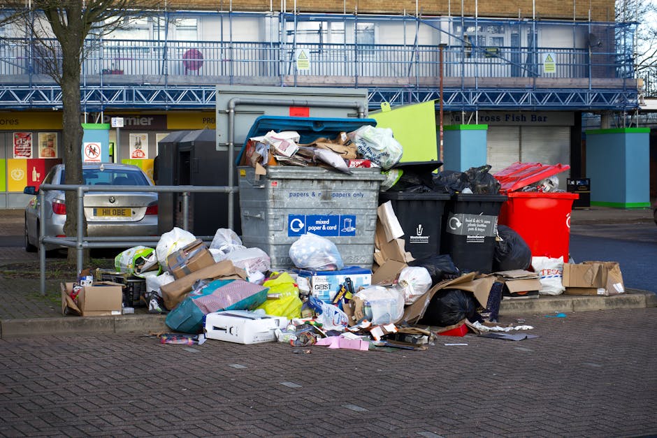 An outdoor scene showing multiple overflowing rubbish bins and bags situated on a paved urban sidewalk near a parking area. The central focus is a large grey mixed waste bin with its lid partially open, revealing crumpled papers, cardboard, and plastic packaging inside. To the right, a black bin contains black plastic garbage bags, and a bright red bin is also visible nearby. Surrounding the bins are scattered cardboard boxes, plastic bags of various colors, and loose waste items, including paper, packaging, and small discarded objects. Behind the waste, there are parked cars, with a metallic grey vehicle positioned on the left side of the image, and a tree with bare branches standing to the left of the bins, suggesting a street scene in a commercial or residential area. In the background, there are storefronts and a building under construction covered with blue scaffolding, with visible signage and storefront windows. The scene reflects an area where waste management and rubbish collection are currently unmanaged, highlighting the importance of proper rubbish disposal practices supported by independent waste clearance services such as those offered by Waste Clearance Barnet.